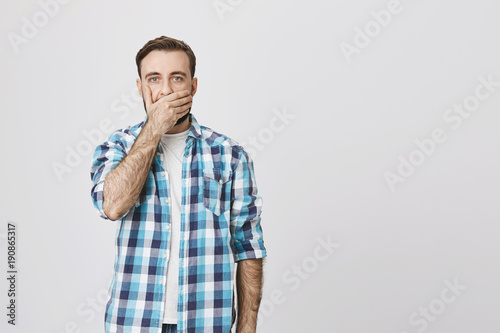 Obraz na plátně Studio shot of terrifired adult man in checked shirt, covering mouth with hand and who is about to cry, against gray wall