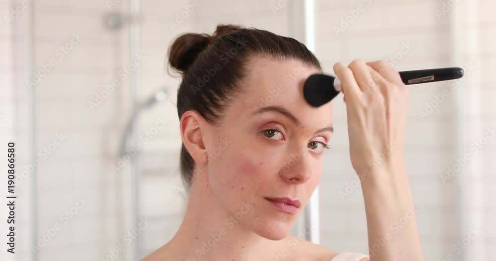 Beauty woman applying makeup. Beautiful girl looking in the mirror and applying cosmetic with a big brush. Girl gets blush on the cheekbones. Powder, rouge
