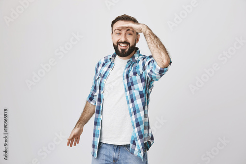 Portrait of cheerful young adult looking far away with hand on his forehead, smiling broadly while standing over gray background. Dad plays hide and seek with his daughter in park