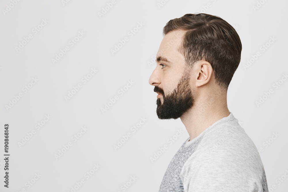 Fototapeta premium Profile portrait of handsome bearded adult european man with trendy haircut, standing over gray background. Hairdresser makes photo of new haircut he did for visitor to post it on site