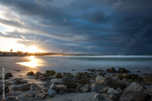Sonnenuntergang am Strand mit Steinen, Palmen und dunklen Wolken in Kuba
