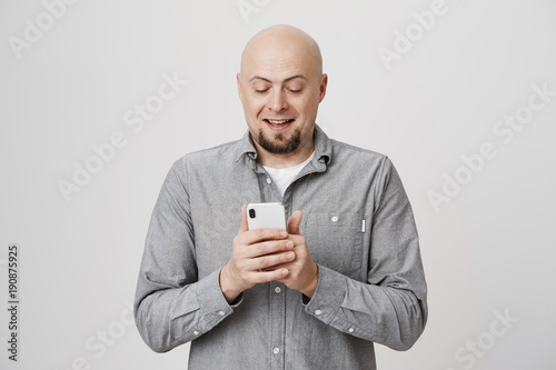 Portrait of charming bald caucasian guy holding smartphone while messaging or browsing, exprassing positive emotions and standing over gray background. Spouse sends wife funny picture of their cat © Cookie Studio