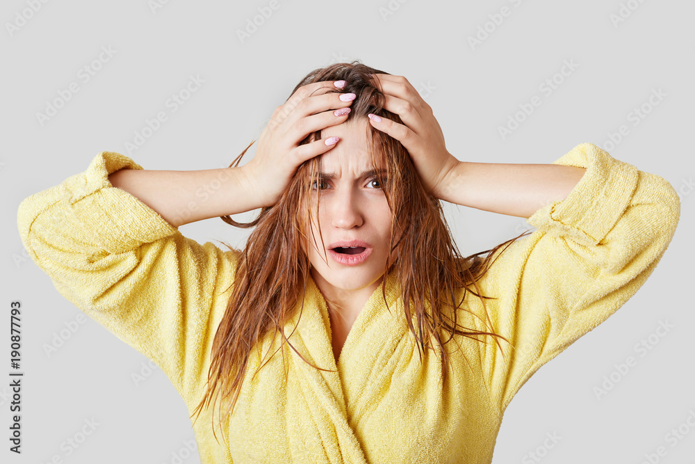 Fototapeta premium Frustrated female touches head, has wet hair and disorder, doesn`t know what to wear and which hairstyle make for meeting, dressed in casual domestic clothing, isolated over white background