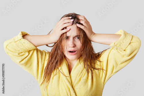 Frustrated female touches head, has wet hair and disorder, doesn`t know what to wear and which hairstyle make for meeting, dressed in casual domestic clothing, isolated over white background