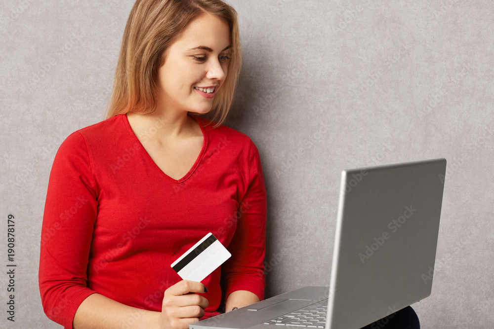 Smiling female buys good in internet store, uses credit card and laptop computer, does online shopping, isolated over grey background. Cute young woman verifies her bank account on notebook Smiling female buys good in internet store, uses credit card and laptop computer, does online shopping, isolated over grey background. Cute young woman verifies her bank account on notebook