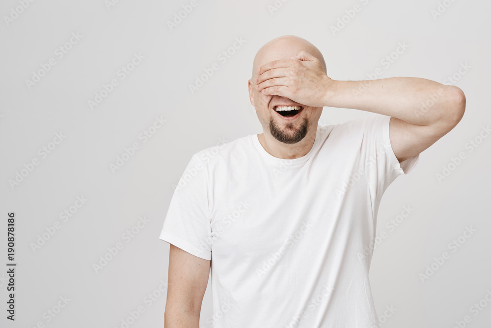 Astonished and excited bald bearded man with broad smile covering eyes with hand and awaiting for something positive to happen, standing over gray background. Birthday guy waits for his present Astonished and excited bald bearded man with broad smile covering eyes with hand and awaiting for something positive to happen, standing over gray background. Birthday guy waits for his present
