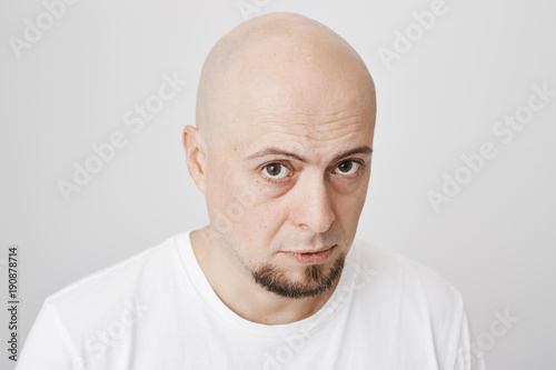 Close-up portrait of handsome bald man with fancy beard, staring up at camera with serious and confident look, standing over gray background. Tired office manager waits till his shift will over.
