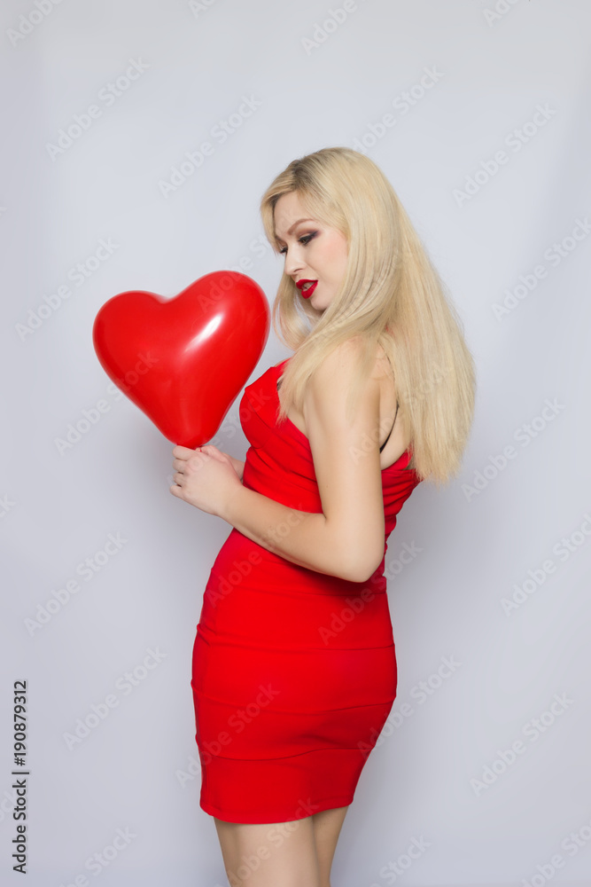 Blonde young woman in elegant red dress. Girl posing on a grey background. Jewelry and hairstyle. With red heart baloon in her hand. Fashion photo Blonde young woman in elegant red dress. Girl posing on a grey background. Jewelry and hairstyle. With red heart baloon in her hand. Fashion photo