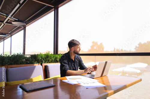 Tired quantity surveyor working at cafe table with diagram and statistic documents. Persistent man dressed in classic black shirt sitting at sofa near green indoor plants. Concept of  preparin photo