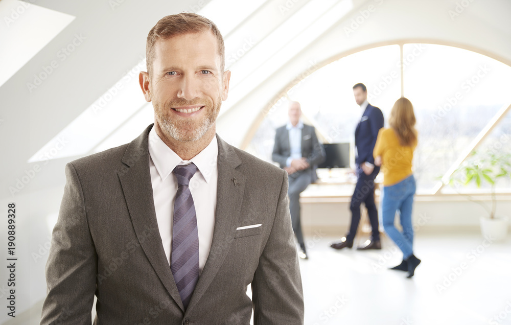 Successful businessman portrait. Senior investment advisor businessman wearing suit while standing at the office and looking at camera. Uncrecognizable business people standing at the background. Successful businessman portrait. Senior investment advisor businessman wearing suit while standing at the office and looking at camera. Uncrecognizable business people standing at the background.