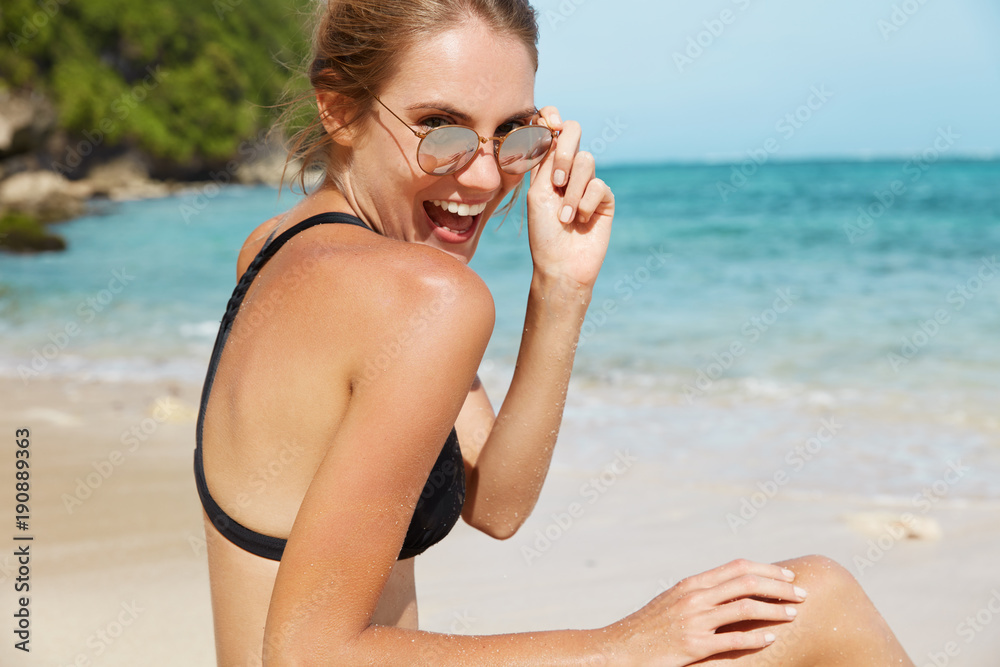 Adorable young female has fun at seashore, glad to pose at camera, looks through sunglasses with joyful expression, sits against wonderful blue marine landscape, has summer vacation in tropics