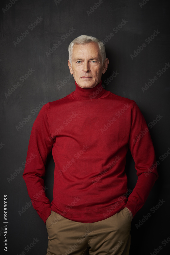 Casual senior man portrait. Handsome elderly man wearing red turtle neck sweater and looking at camera while standing against at dark background. Casual senior man portrait. Handsome elderly man wearing red turtle neck sweater and looking at camera while standing against at dark background.