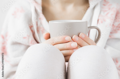 Woman in white soft home sweater holding a mug in hands at home. Enjoying coffee or tea in day off. Morning drink concept.