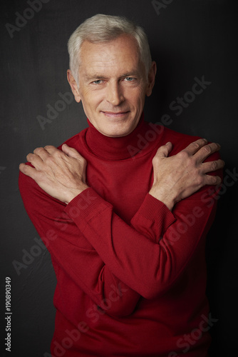 Studio portrait of handsome elderly man wearing turtle neck sweater and looking at camera while standing against at dark background.