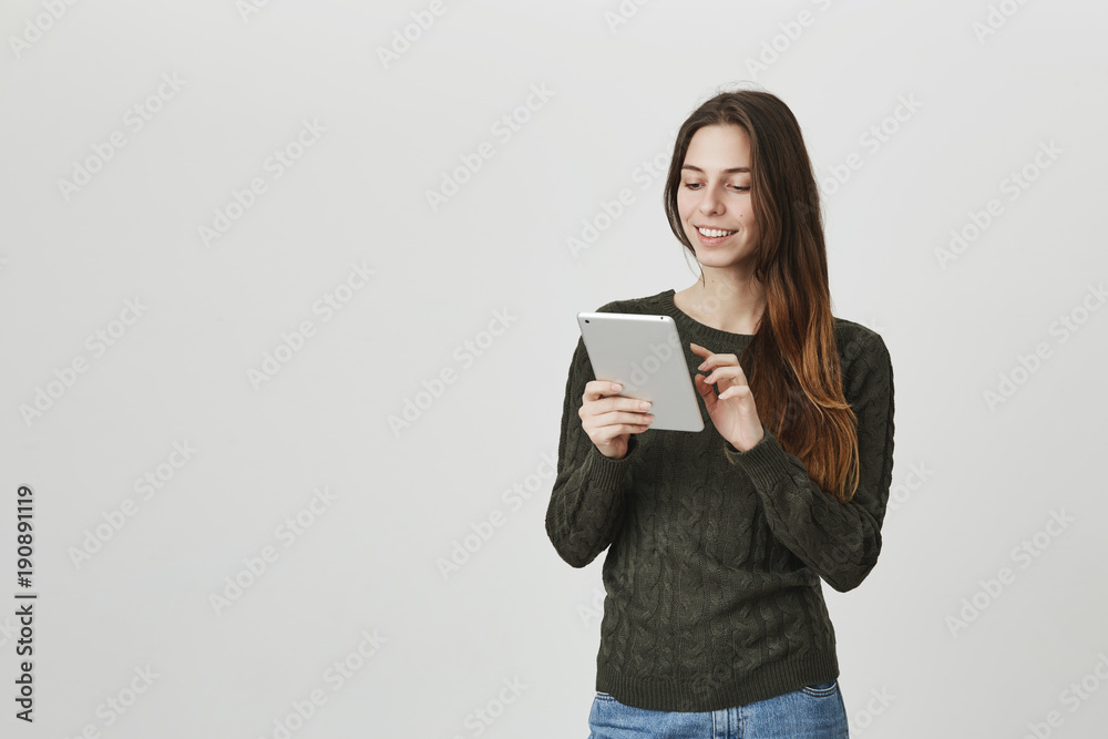 Portrait of attractive young female smiling widely scrolling news in her tablet, wearing casual sweater over white background. Student watches funny and cute video with cats in social network Portrait of attractive young female smiling widely scrolling news in her tablet, wearing casual sweater over white background. Student watches funny and cute video with cats in social network