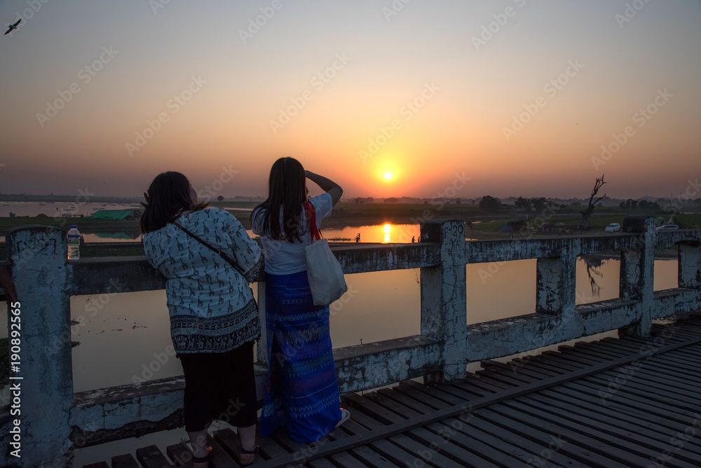 Fototapeta premium Tourist women standing and seeing and shooting beautiful view of sunset at U-Bein wooden bridge in Mandalay, Myanmar. 