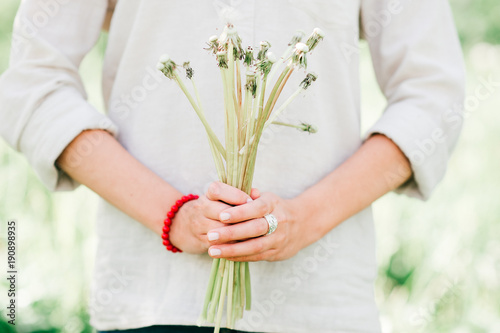 Beuatiful model girl with lovely kind cheerful face holding bouquet of dandelions in hands on emerald abstract background outdoor at nature. Spring flowers. Blooming garden. Joy of life. Sunny day.