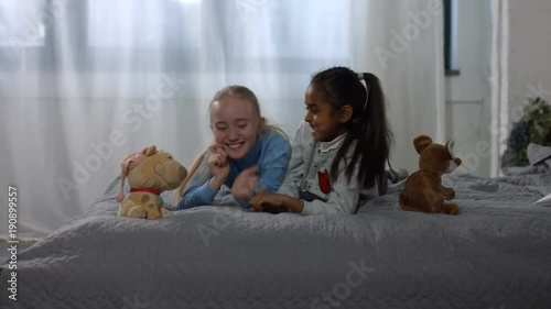 Excited cute multi ethnic preteen girls having fun and playing on the bed at home. Carefree multiracial kids enjoying leisure together ,playing , tickling and laughing in domestic room.