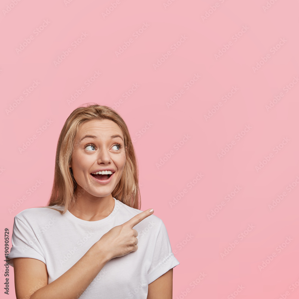 Happy excited female with long light hair indicates with fore finger aside, demonstrates something at blank copy space for your advertisment or hearder. Cute woman in casual t shirt gestures indoor Happy excited female with long light hair indicates with fore finger aside, demonstrates something at blank copy space for your advertisment or hearder. Cute woman in casual t shirt gestures indoor