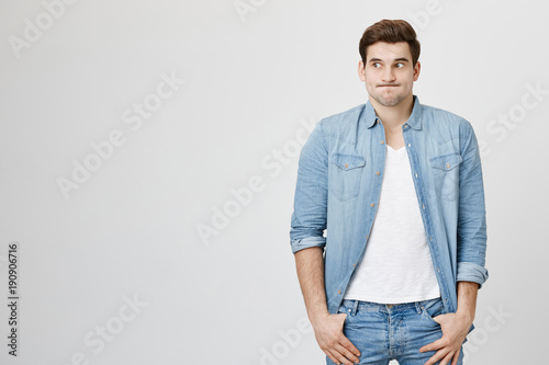 Studio shot of emotional clueless man with trendy haircut in denim shirt over white t-shirt having confused puzzled look, pouting cheeks, shrugging shoulders as he doesn't know reason of accident