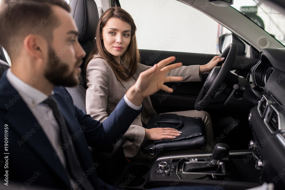 Portrait of beautiful young woman sitting behind wheel of brand new luxury car and talking to salesman while choosing new transport