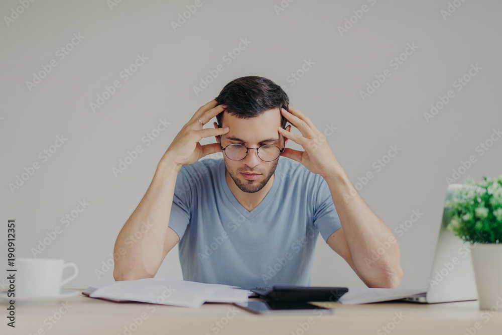 Young European male dressed casually, has debt problems, not able to pay out all debts, studies papers from bank, has frustrated look, tries to manage finances, calculates loan with calculator Young European male dressed casually, has debt problems, not able to pay out all debts, studies papers from bank, has frustrated look, tries to manage finances, calculates loan with calculator