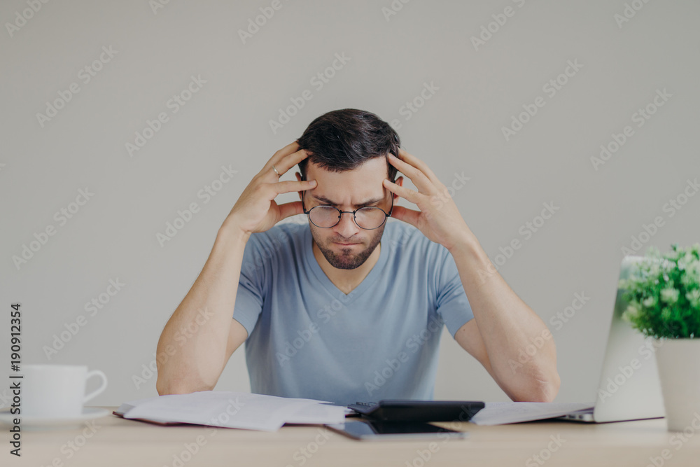 Brunet young male faces financial problems, not able to pay out mortagage, has desperate expression, makes calculations with calculator, isolated over grey background, uses modern technologies Brunet young male faces financial problems, not able to pay out mortagage, has desperate expression, makes calculations with calculator, isolated over grey background, uses modern technologies