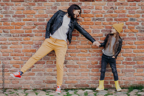 Small fashionable girl and her mother have fun together, keep hands together, pose against brick wall. Stylish young mum and daughter play outdoor. Relationships, good mood and fashion concept