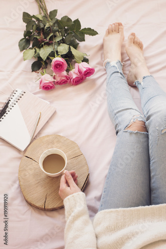 Young Caucasian woman in bed early in the morning, wearing distressed jeans and a white sweater and holds a cup of coffee. Roses and notebooks around. Cozy blogger mornings