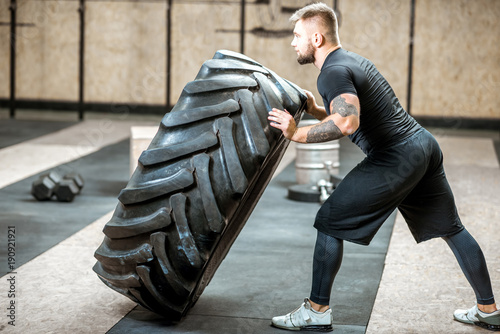 Athletic man in black sportswear pushing a big tire training in the gym