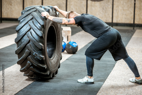 Athletic man in black sportswear pushing a big tire training in the gym