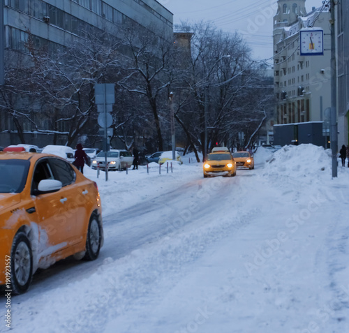 A yellow taxi rides along the winter road in the city after a snowfall. Twilight. A lot of snow. Drifts.