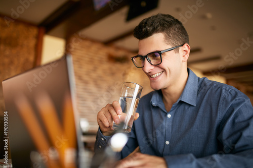 Fotografie Smiling caucasian man with glass of water in hand works with laptop