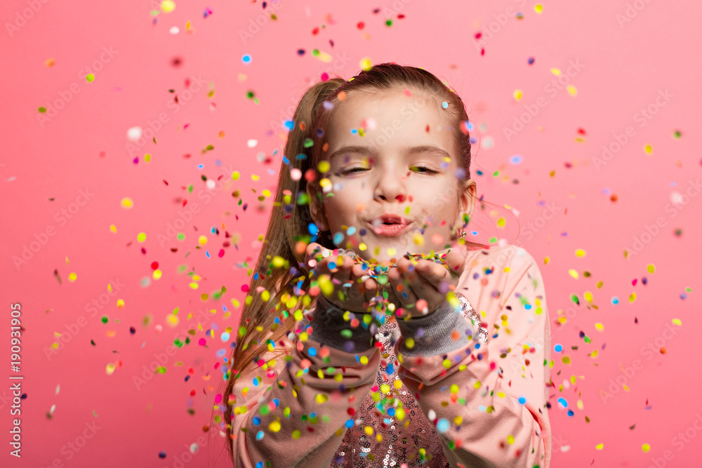 Happy girl celebrating on a pink background. Blows up multicolored confetti