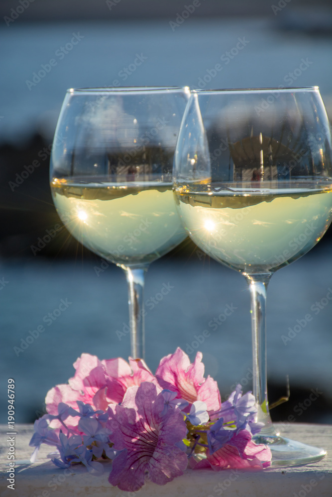 Obraz premium Two glasses with cold white wine served outside on terrace with pink tropical flowers, luxury resort with sea view, romantic vacation