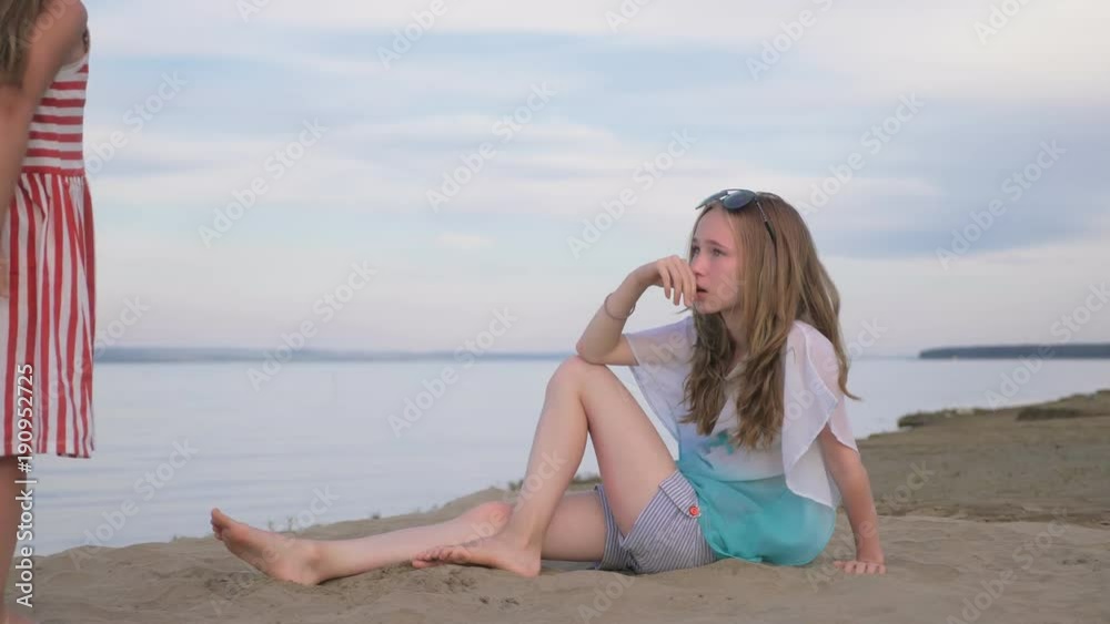 Two teenage children are sitting on the beach. They wear sunglasses, they play with sand. Kid indulge. Sisters communicate with each other. Two teenage children are sitting on the beach. They wear sunglasses, they play with sand. Kid indulge. Sisters communicate with each other.