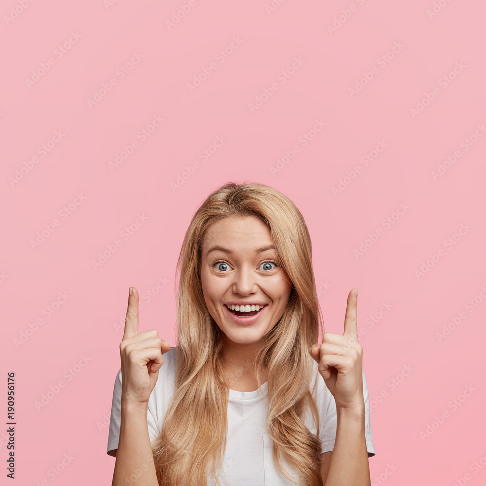 Isolated shot of pretty blonde female with broad smile, appealing look, indicates with fore fingers up, glad to advertize something, blank copy space for your promotional text or advertisment Isolated shot of pretty blonde female with broad smile, appealing look, indicates with fore fingers up, glad to advertize something, blank copy space for your promotional text or advertisment