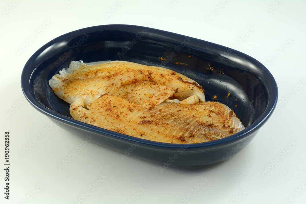 Fresh raw tilapia fish fillets prepared for cooking with spicy rub of chili powder, garlic and cumin in blue oval dish on white wooden table Fresh raw tilapia fish fillets prepared for cooking with spicy rub of chili powder, garlic and cumin in blue oval dish on white wooden table