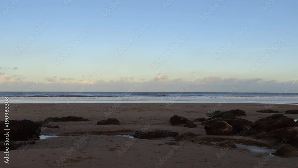 Panning shot of Scarborough, England beach at sunset, man wearing jacket with hood is walking his dog, castle ruins on the hill Panning shot of Scarborough, England beach at sunset, man wearing jacket with hood is walking his dog, castle ruins on the hill