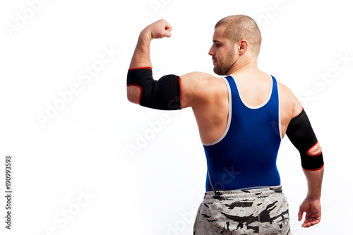 Young athletic man in blue tights and black elbow stands with back to camera and shows biceps on white isolated background