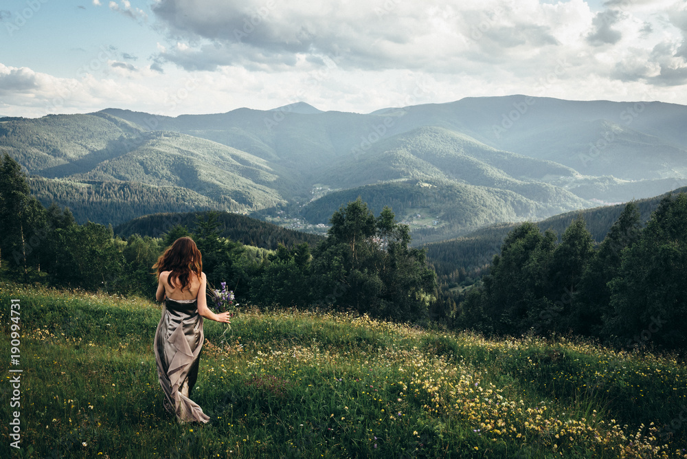 A beautiful girl in a dress and a bouquet of flowers in her hands, on a green glade dancing among the peaks of the mountains