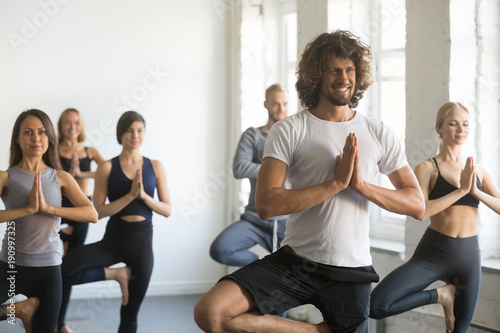 Group of young sporty people practicing yoga lesson with instructor, standing in Vrksasana exercise, Tree pose, working out, indoor close up, students training in club, studio © fizkes