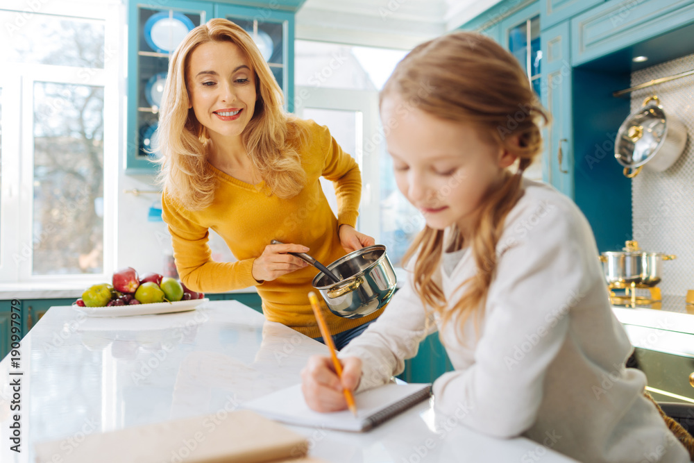 I cherish you. Beautiful alert blond young mother smiling and holding a saucepan while looking at her little daughter writing in her notebook