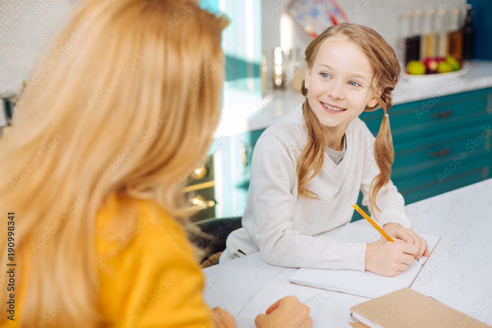 My dear mum. Attractive joyful fair-haired girl smiling and holding a pencil while looking at her mother and sitting at the table My dear mum. Attractive joyful fair-haired girl smiling and holding a pencil while looking at her mother and sitting at the table