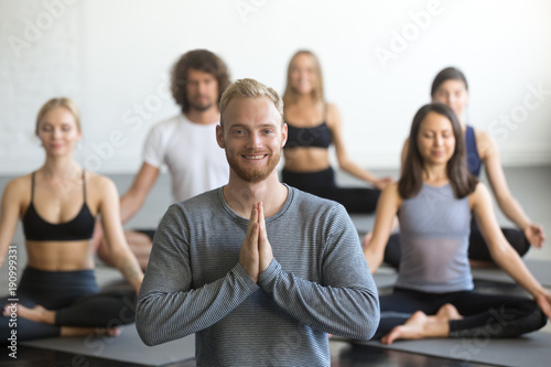 Portrait of young friendly smiling male yoga instructor looking at camera, group of sporty people practicing lotus pose, sitting in Padmasana exercise, working out, students training in club, studio