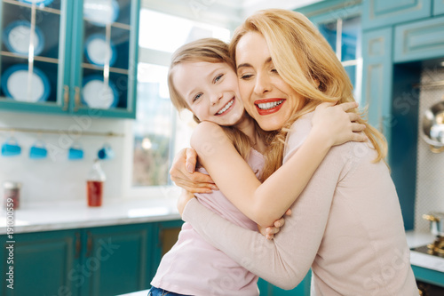 My treasure. Pretty cheerful fair-haired young mother smiling and hugging her daughter and her daughter beaming