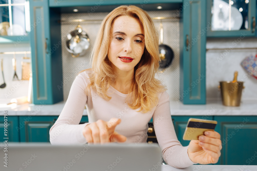 Determined freelancer. Attractive concentrated fair-haired young woman holding a card and working on her laptop while sitting in the kitchen Determined freelancer. Attractive concentrated fair-haired young woman holding a card and working on her laptop while sitting in the kitchen