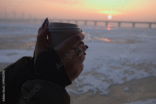 Women's hands hold coffee paper cup. Amazing sunrise on a cool winter morning in the background. River Dnipro, Kyiv, Ukraine