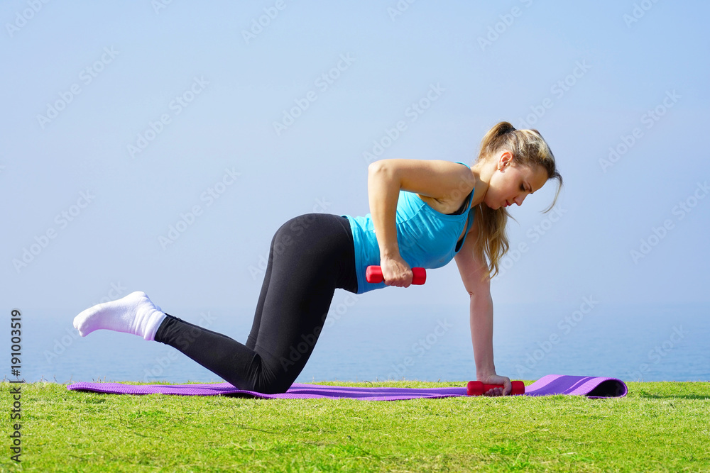 Fototapeta premium Young girl on grass with dumbbell in hand on the background of the sea. Fitness girl doing exercises with weights on the beach under the spring sun. Concept of health and weight loss.