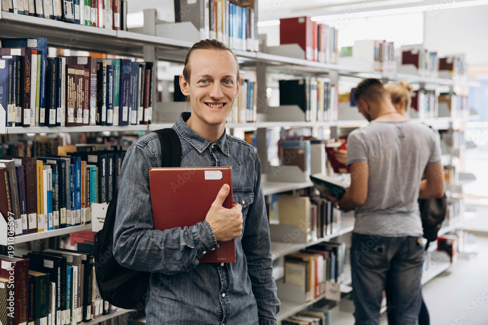 Thoughtful boy looks like a student standing with book in the library of a university Thoughtful boy looks like a student standing with book in the library of a university